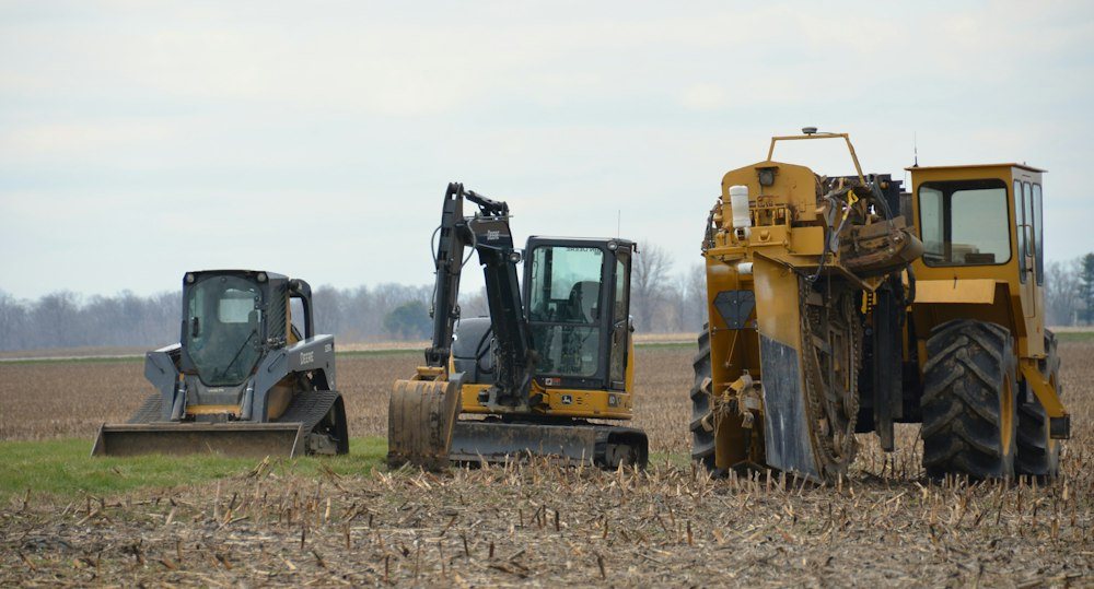 A compact track loader parked at the edge of a field beside a hardwood tree line